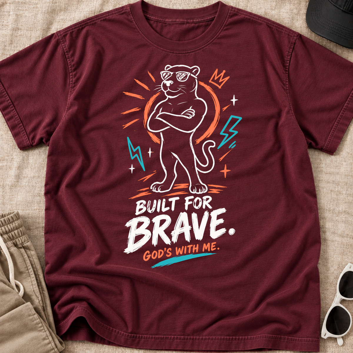 A maroon short-sleeve graphic T-shirt laid flat on a beige fabric surface, with a stylized panther illustration and the text 'BUILT FOR BRAVE. GOD'S WITH ME.