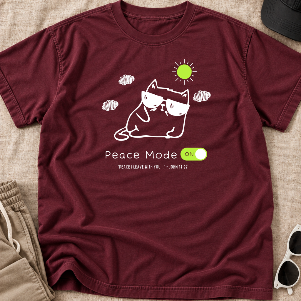 A maroon crewneck t-shirt laid flat on a beige fabric background, featuring a white line art graphic of a cat in a peace pose with sunglasses, a green sun, and the text 'Peace Mode ON' with 'PEACE LEAVE WITH YOU - JOHN 14:27'.