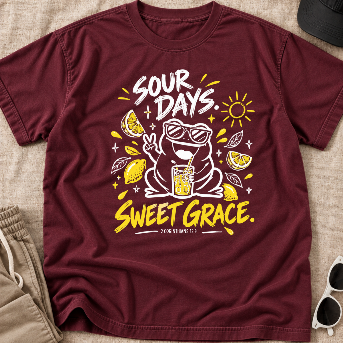 A maroon short-sleeve graphic T-shirt laid flat on a beige fabric surface, with part of a khaki pair of shorts, a black cap, and white sunglasses visible around it. The shirt graphic shows a white cartoon frog wearing sunglasses and making a peace sign, surrounded by yellow lemon slices, a drink, and the text 'SOUR DAYS.