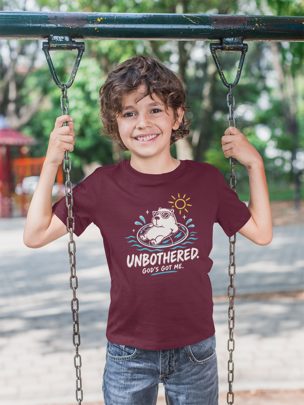 A smiling young boy stands on a playground swing holding the chains, wearing a maroon Christian graphic t-shirt with a cartoon capybara and the text 'UNBOTHERED. GOD'S got ME.