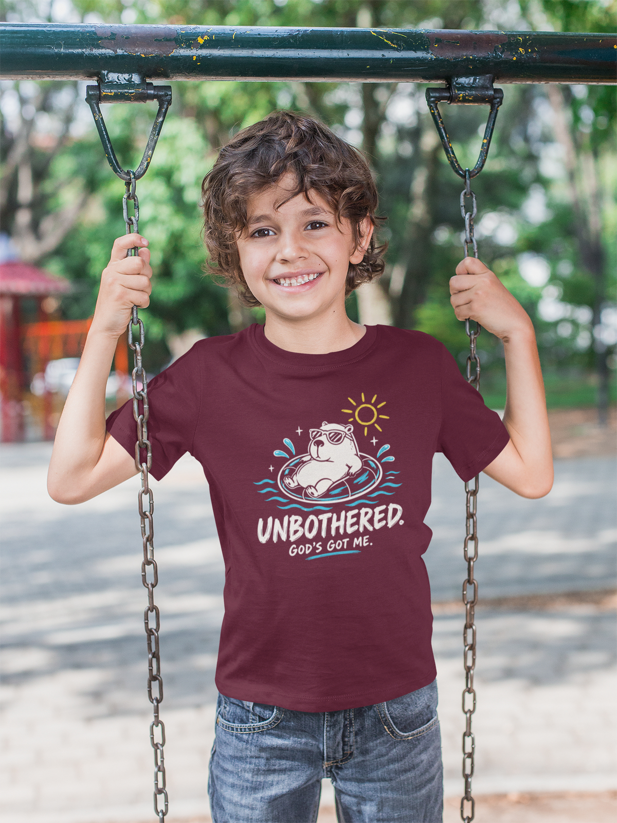 A smiling young boy stands on a playground swing holding the chains, wearing a maroon Christian graphic t-shirt with a cartoon capybara and the text 'UNBOTHERED. GOD'S got ME.
