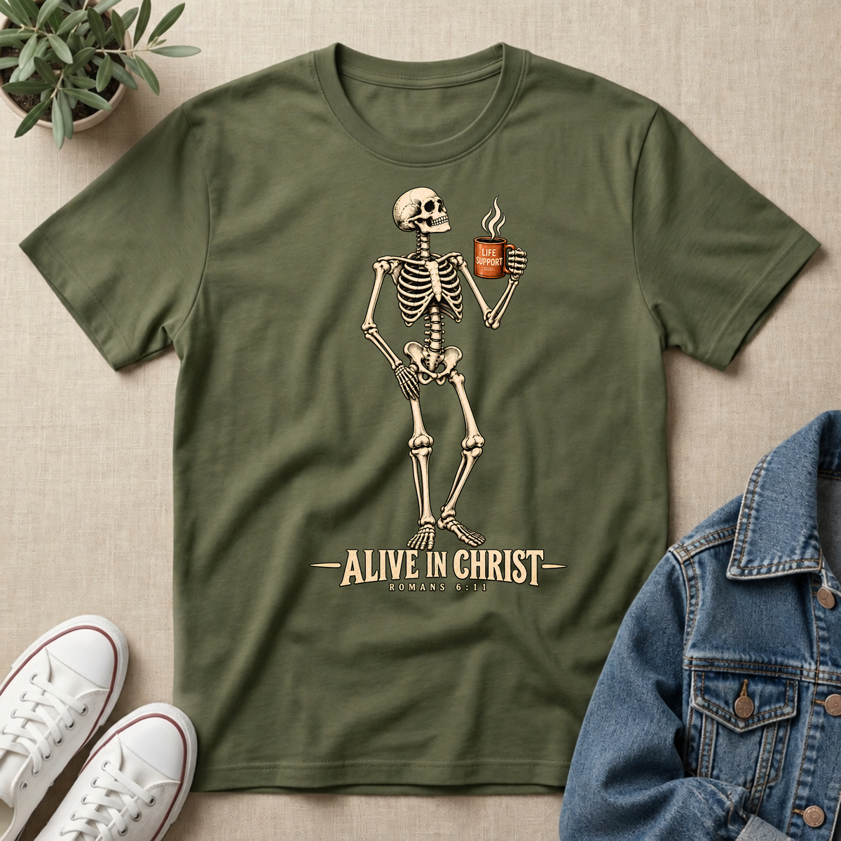 A military green short-sleeve t-shirt with a graphic of a skeleton holding a cup, with the text 'ALIVE IN CHRIST ROMANS 8:11' printed below. The t-shirt is displayed in a flat-lay arrangement on a beige textured background, surrounded by a small potted plant, white sneakers, and a blue denim jacket.