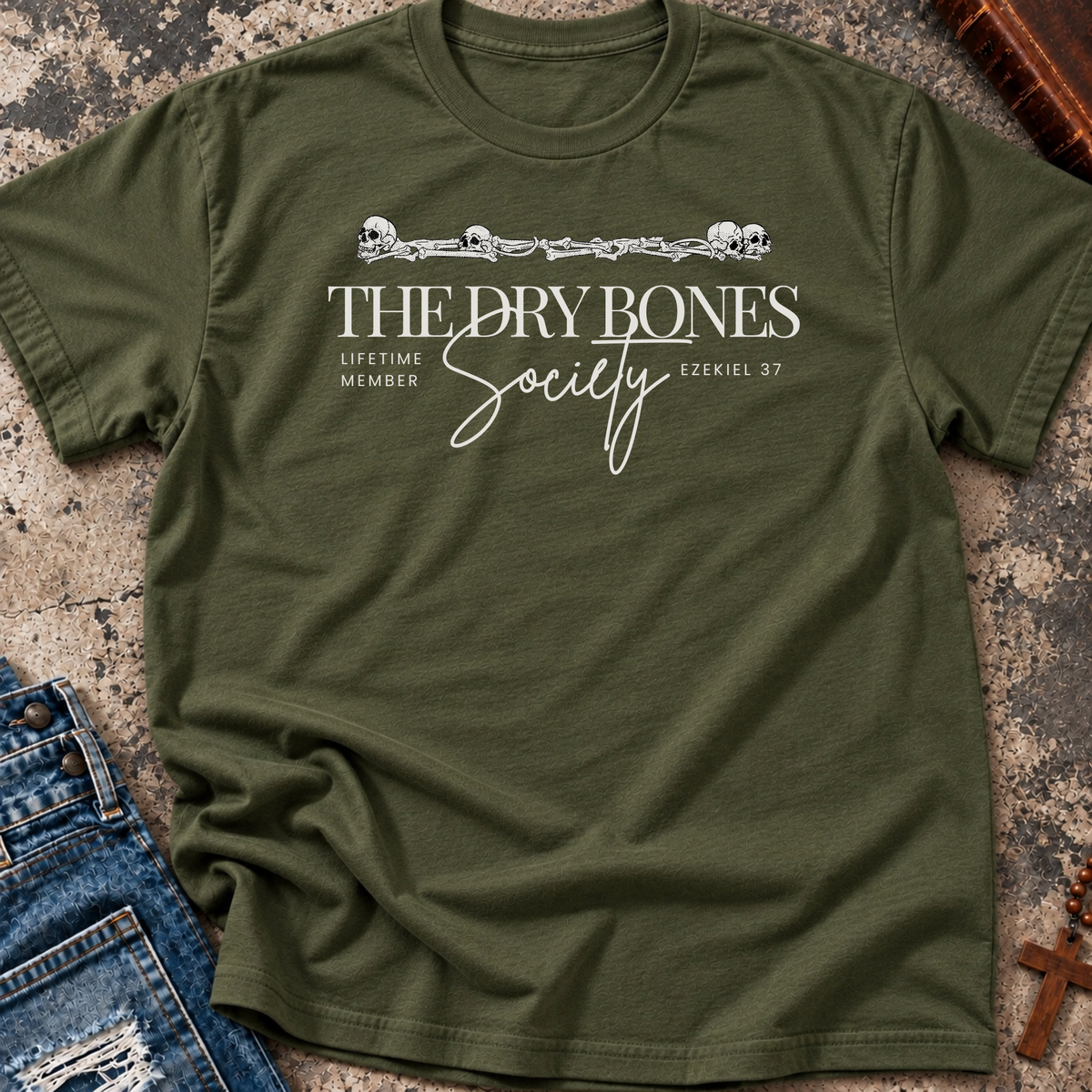 A military green Christian graphic t-shirt by SeedSowerGraphics laid flat on a textured stone surface, styled with a pair of blue jeans on the left and a wooden cross in the bottom right corner. The t-shirt features a white printed graphic with skulls, a barbed wire design, and the text 'THE DRY BONES Society' and 'LIFETIME MEMBER EZEKIEL 37'.