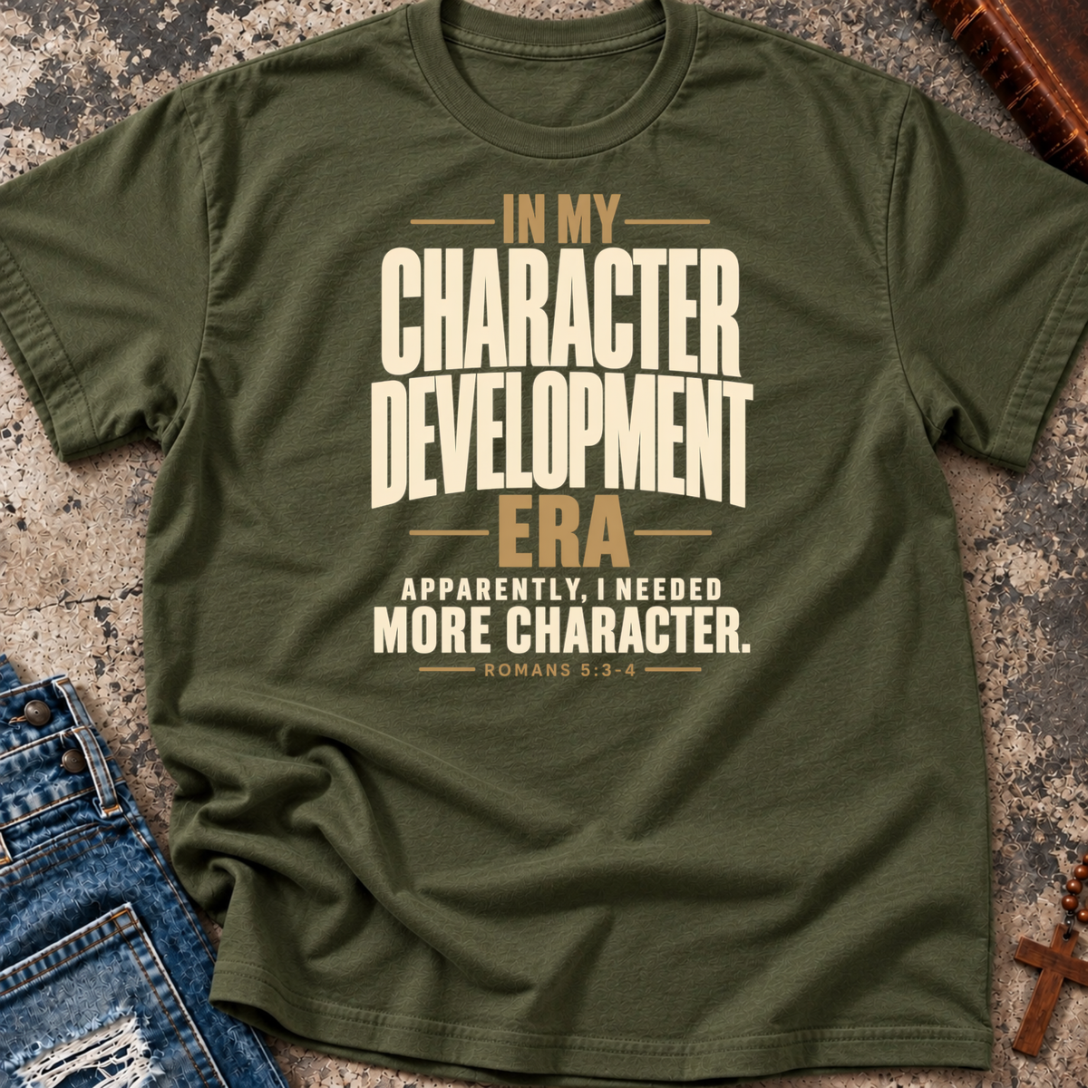 A military green Christian graphic t-shirt by SeedSowerGraphics laid flat on a textured stone surface, styled with blue jeans and a wooden cross. The t-shirt features a beige and brown typographic print that reads 'IN MY CHARACTER DEVELOPMENT ERA APPARENTLY, I NEEDED MORE CHARACTER.