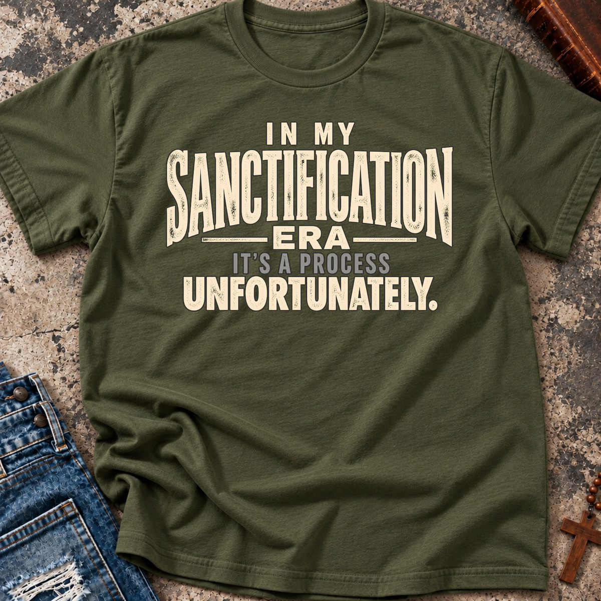 A military green graphic t-shirt laid flat on a textured stone surface, styled with a pair of blue jeans and a wooden cross. The t-shirt features a distressed cream-colored text graphic that reads: 'IN MY SANCTIFICATION ERA IT'S A PROCESS UNFORTUNATELY.