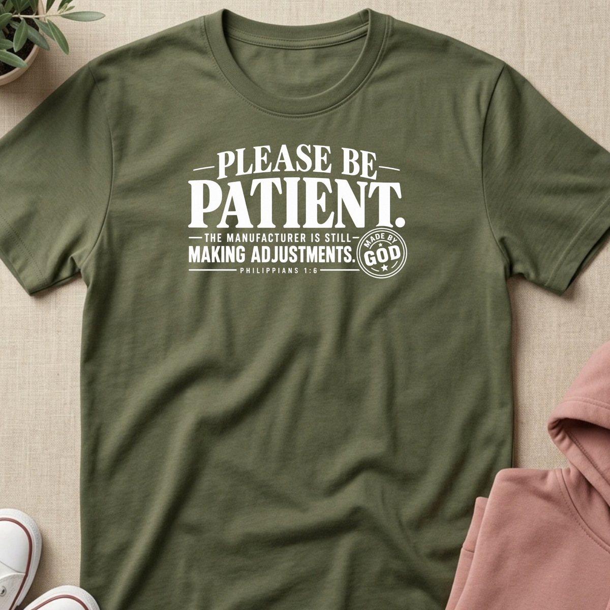 An olive green short-sleeve t-shirt with a white graphic print that reads 'PLEASE BE PATIENT. THE MANUFACTURER IS STILL MAKING ADJUSTMENTS.
