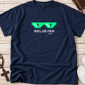 A navy blue short-sleeve t-shirt laid flat on a light wood surface, featuring a green pixelated sunglasses graphic with white crosses and the word 'BELIEVER' and 'JON 3:16' printed below. A brown cross, rosary, book, and white sneakers are partially visible around the edges as styling props.