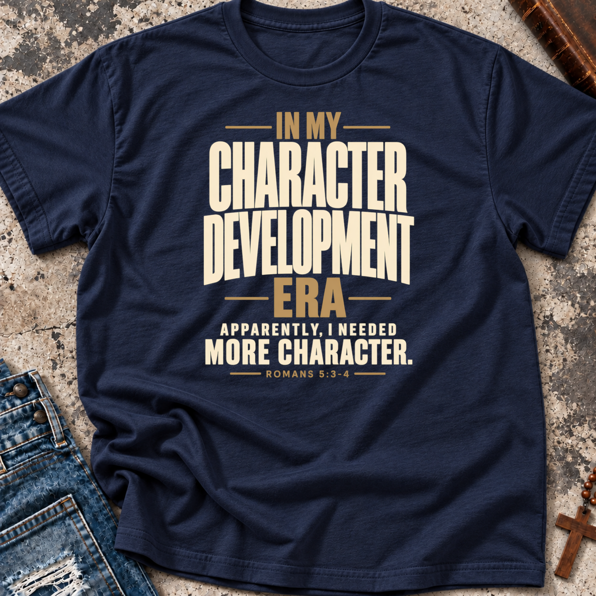 A navy blue Christian graphic t-shirt by SeedSowerGraphics laid flat on a textured stone surface, styled with a pair of distressed blue jeans on the left and a brown leather cross on the right. The t-shirt features a beige and gold typographic design that reads 'IN MY CHARACTER DEVELOPMENT ERA APPARENTLY, I NEEDED MORE CHARACTER.