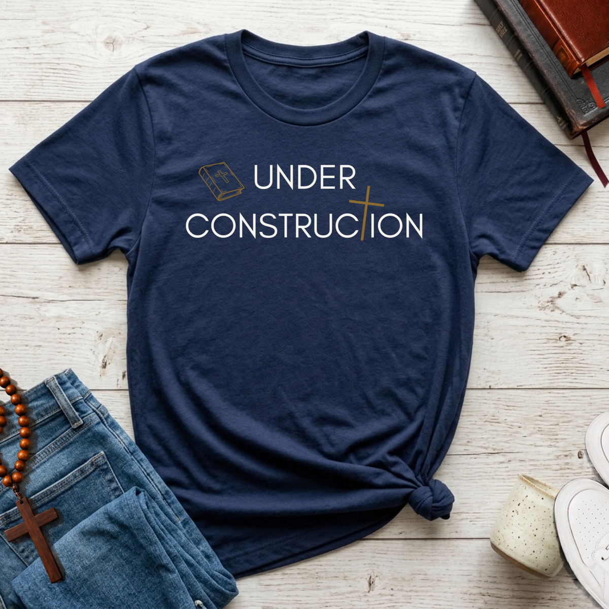A navy blue graphic t-shirt with the words 'UNDER CONSTRUCTION' printed in white, styled in a flat lay on a light wood background with jeans, a rosary, and books.