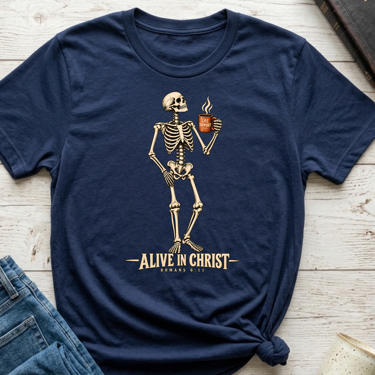 A navy blue short-sleeve t-shirt with a graphic of a standing skeleton holding a coffee mug and the text 'ALIVE IN CHRIST ROMANS 8:11' printed below. The t-shirt is styled flat on a light wood background with a knot at the bottom hem, alongside a pair of blue jeans.
