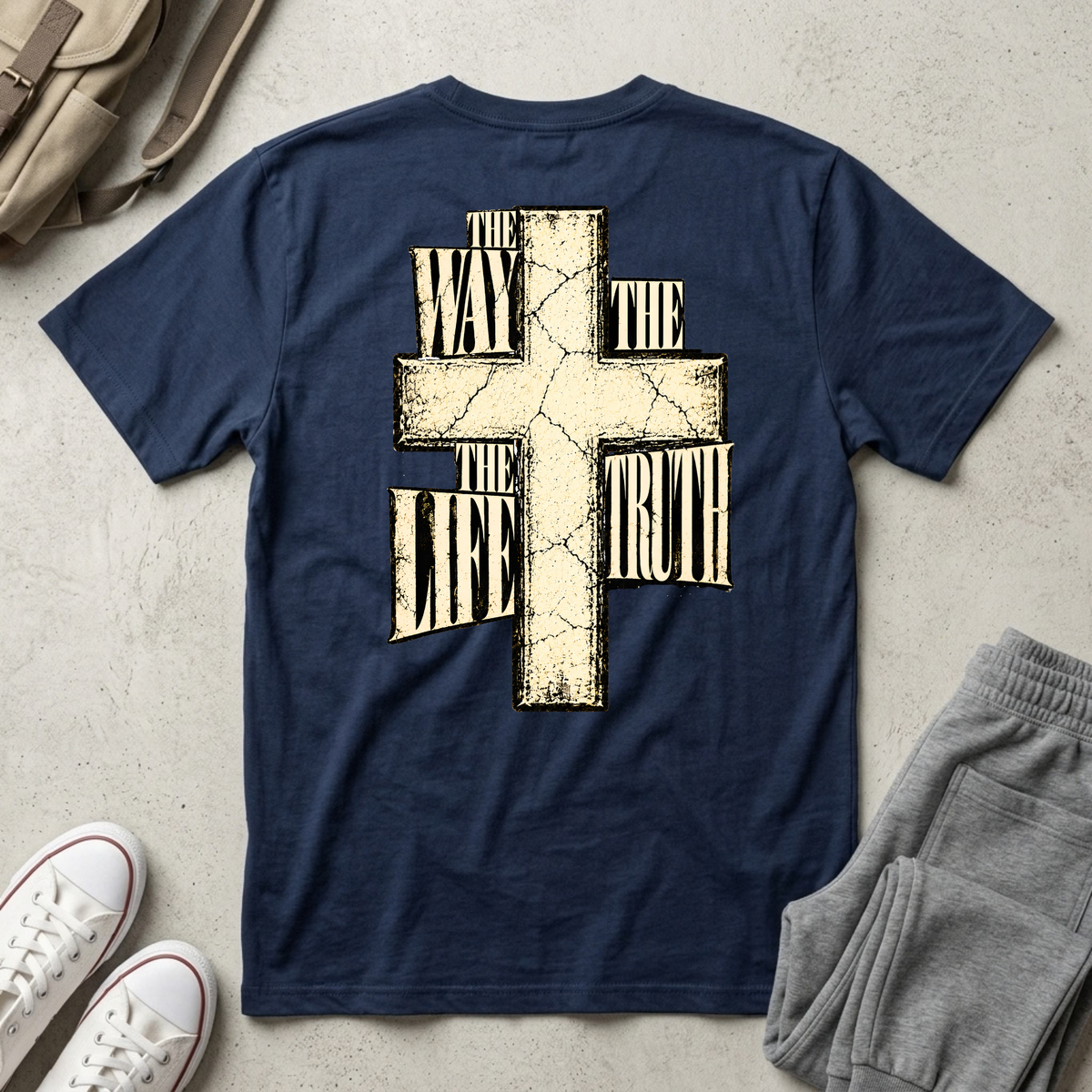 A navy blue short-sleeve t-shirt with a large distressed beige cross graphic and the text 'THE WAY THE LIFE THE TRUTH' printed on the back, styled in a flat-lay with gray sweatpants, white sneakers, and a tan backpack on a light concrete floor.