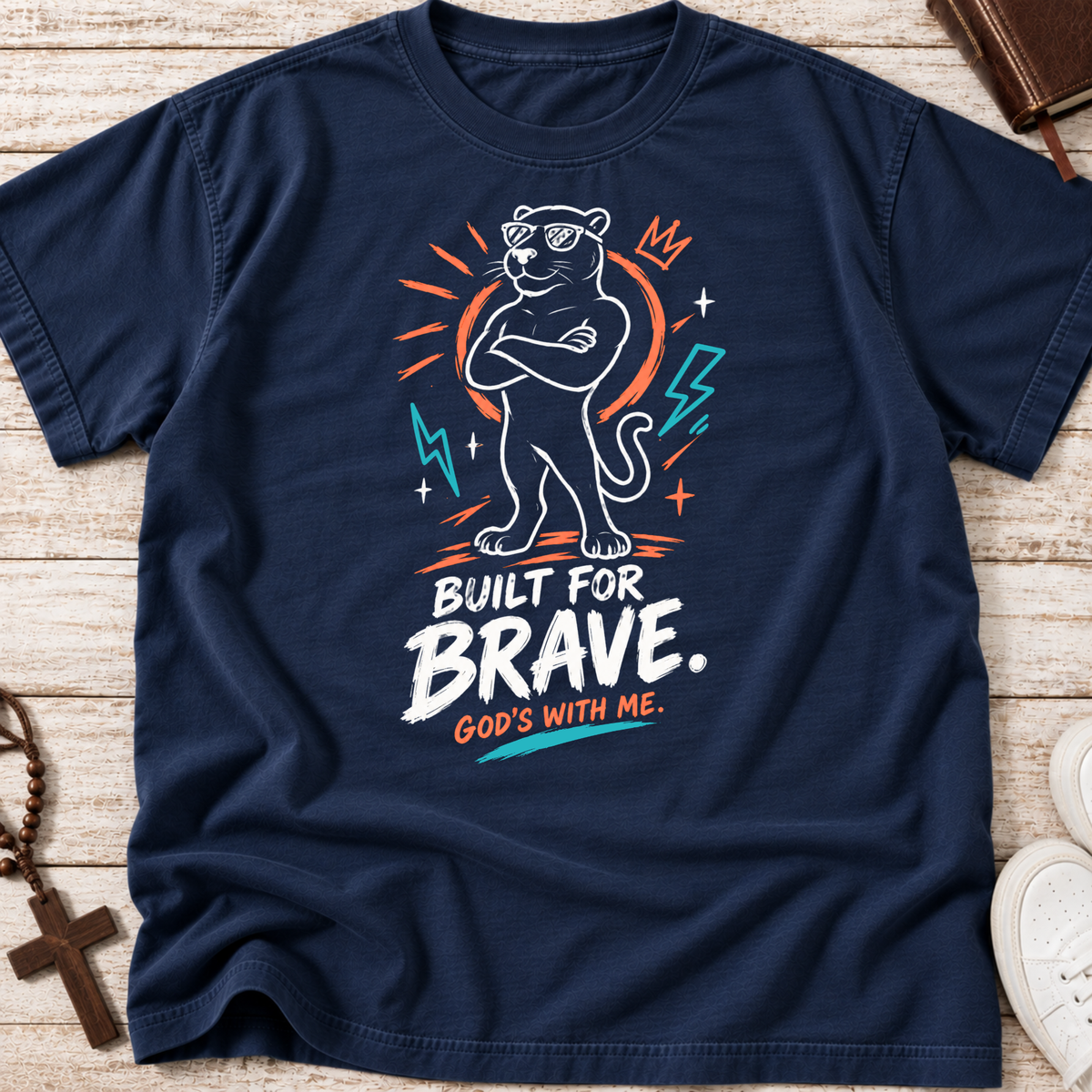 A navy blue graphic t-shirt laid flat on a light wood background. The shirt features a stylized panther wearing sunglasses with its arms crossed, surrounded by orange and teal lightning bolts and stars, with the text 'BUILT FOR BRAVE.