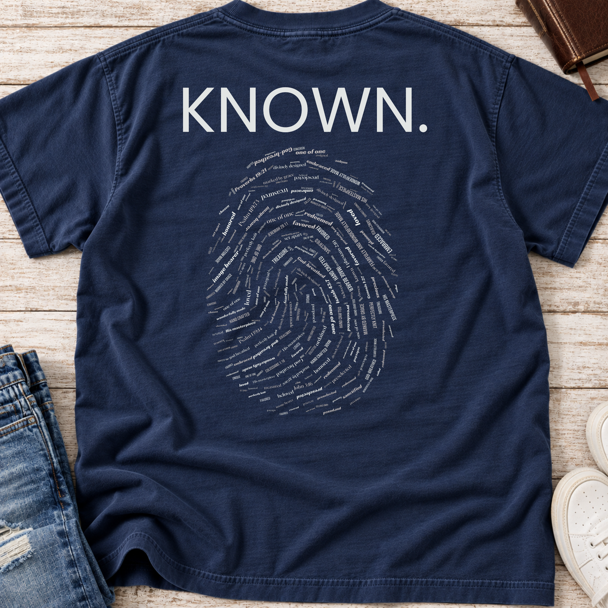 A navy blue short-sleeve T-shirt laid flat on a textured beige surface, with the back printed in white text reading "KNOWN." above a large fingerprint made of small words.