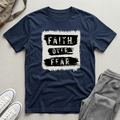 A navy blue short-sleeve t-shirt with a distressed white and black graphic reading 'FAITH OVER FEAR', styled in a flat lay with gray sweatpants, white sneakers, and a beige tote bag on a light gray surface.