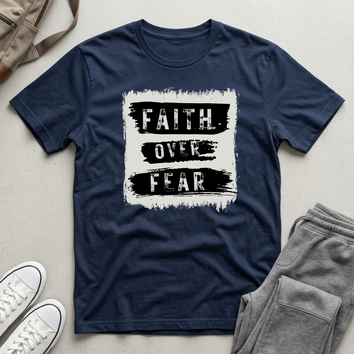 A navy blue short-sleeve t-shirt with a distressed white and black graphic reading 'FAITH OVER FEAR', styled in a flat lay with gray sweatpants, white sneakers, and a beige tote bag on a light gray surface.