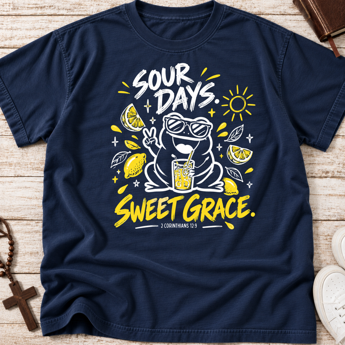 A navy blue graphic t-shirt laid flat on a light wood background. The shirt features a white and yellow cartoon frog wearing sunglasses, surrounded by lemons, a drink, and the text 'SOUR DAYS.