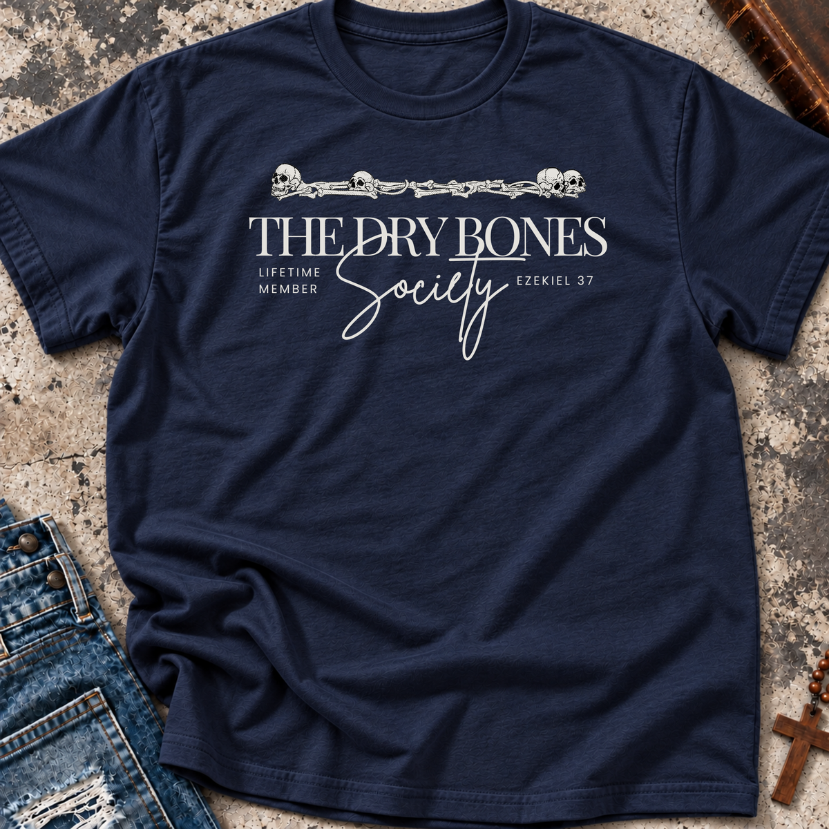A navy blue short-sleeve Christian graphic t-shirt by SeedSowerGraphics with a white graphic print reading 'THE DRY BONES Society' and 'LIFETIME MEWDER' with 'EZEKIEL 37' below, styled flat on a textured gray surface with jeans and a wooden cross partially visible.