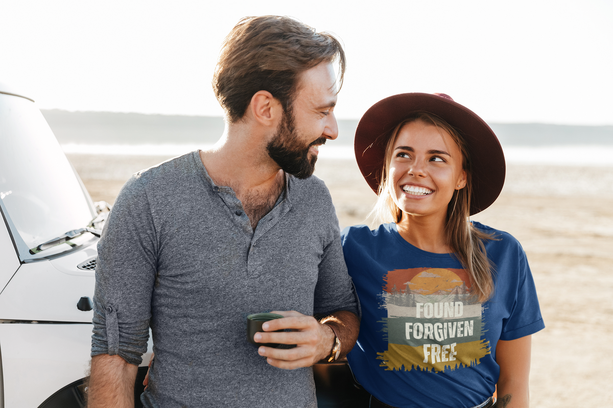 A smiling woman in a wide-brimmed brown hat and blue Christian graphic T-shirt stands beside a man in a gray henley shirt next to a white vehicle in a bright outdoor desert setting. The T-shirt features a retro sunset graphic with the text 'FOUND FORGIVEN FREE'.
