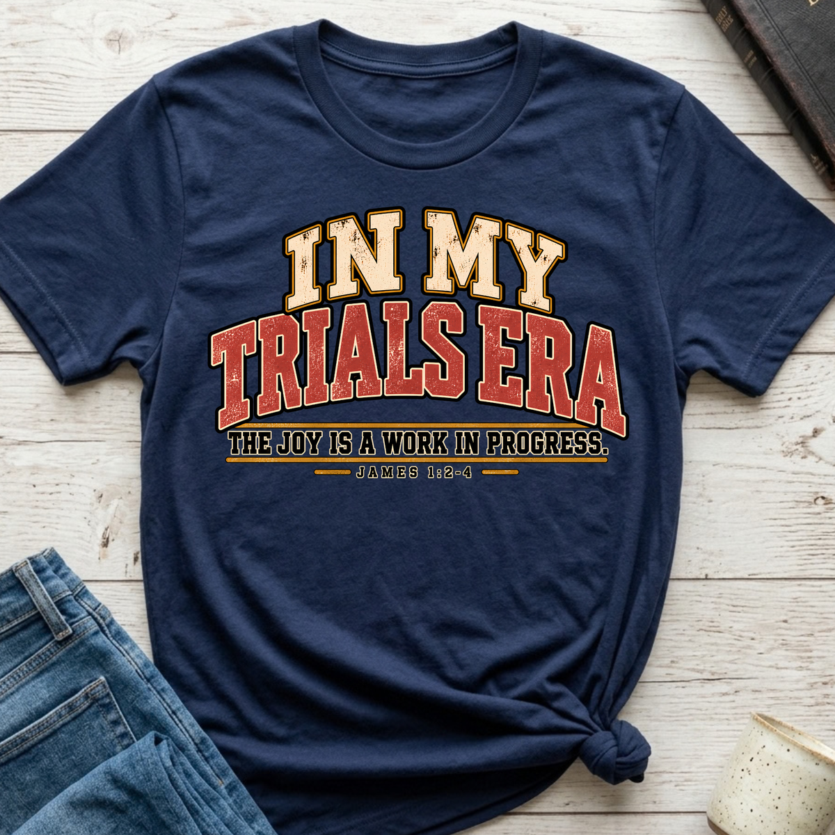 navy christian t-shirt by SeedSowerGraphics  laid out amongst props with typography that says "in my trials era' the joy is a work in progress. James 1:2-4 in cream, red, and black lettering