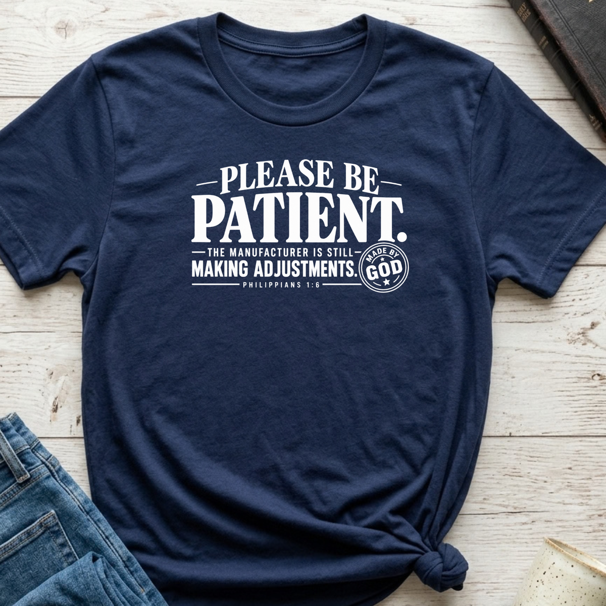 A navy blue graphic t-shirt with white text reading 'PLEASE BE PATIENT. THE MANUFACTURER IS STILL MAKING ADJUSTMENTS.