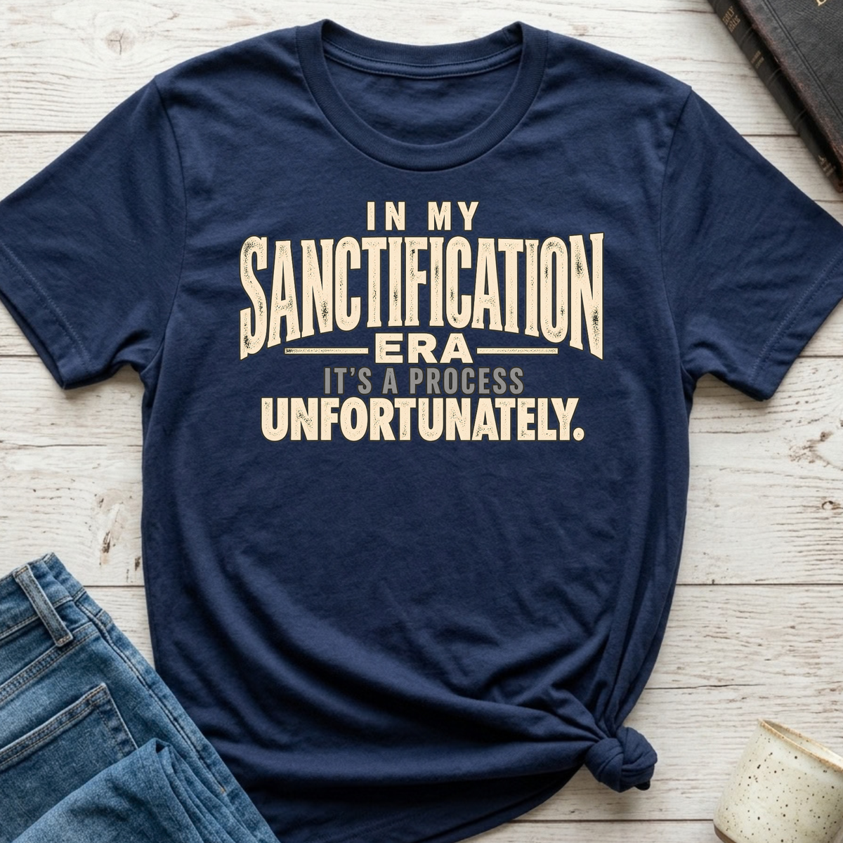 A navy blue graphic t-shirt with a distressed cream-colored text print reading 'IN MY SANCTIFICATION ERA IT'S A PROCESS UNFORTUNATELY.' styled in a flat-lay on a light wood background with jeans and a mug partially visible.