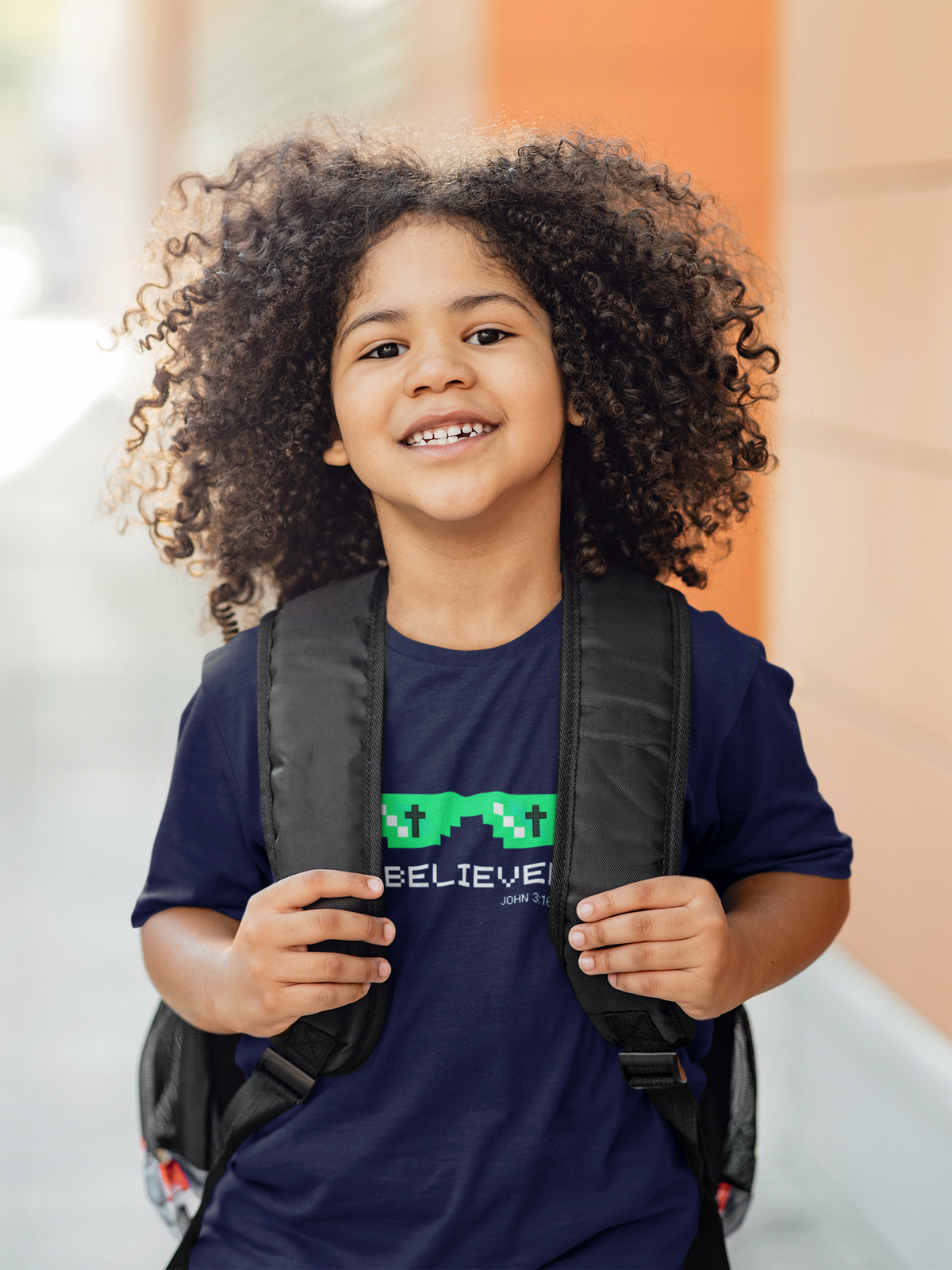A smiling young girl with voluminous curly hair is shown from the chest up, wearing a navy blue youth Christian graphic t-shirt and a black backpack. The t-shirt features a green pixelated design above the word 'BELIEVER".