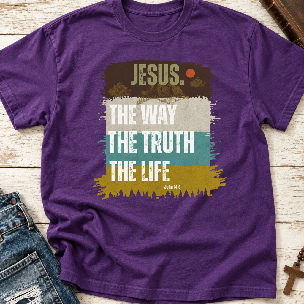 A purple short-sleeve graphic T-shirt laid flat on a whitewashed wood surface, styled with blue jeans at the lower left and a small cross at the lower right. The shirt has a large front print reading “JESUS.