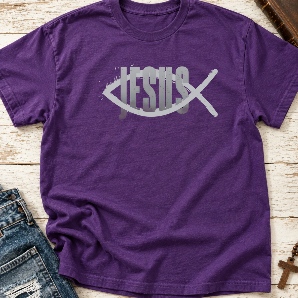A purple short-sleeve t-shirt with a graphic reading 'JESUS' in white and gray, overlaid with a white Ichthys symbol, laid flat on a light wood background with jeans and a cross necklace partially visible.