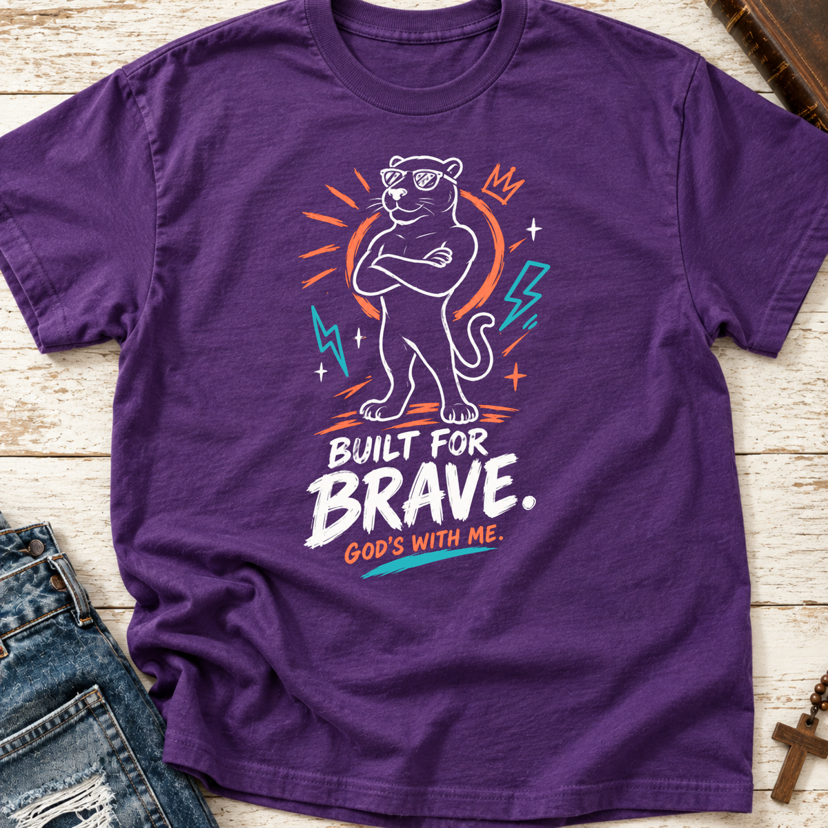 A purple short-sleeve graphic T-shirt laid flat on a light wood surface, styled with part of a blue denim jacket at the lower left. The shirt features a white panther illustration with sunglasses, orange and teal lightning-bolt accents, and the text 'BUILT FOR BRAVE.