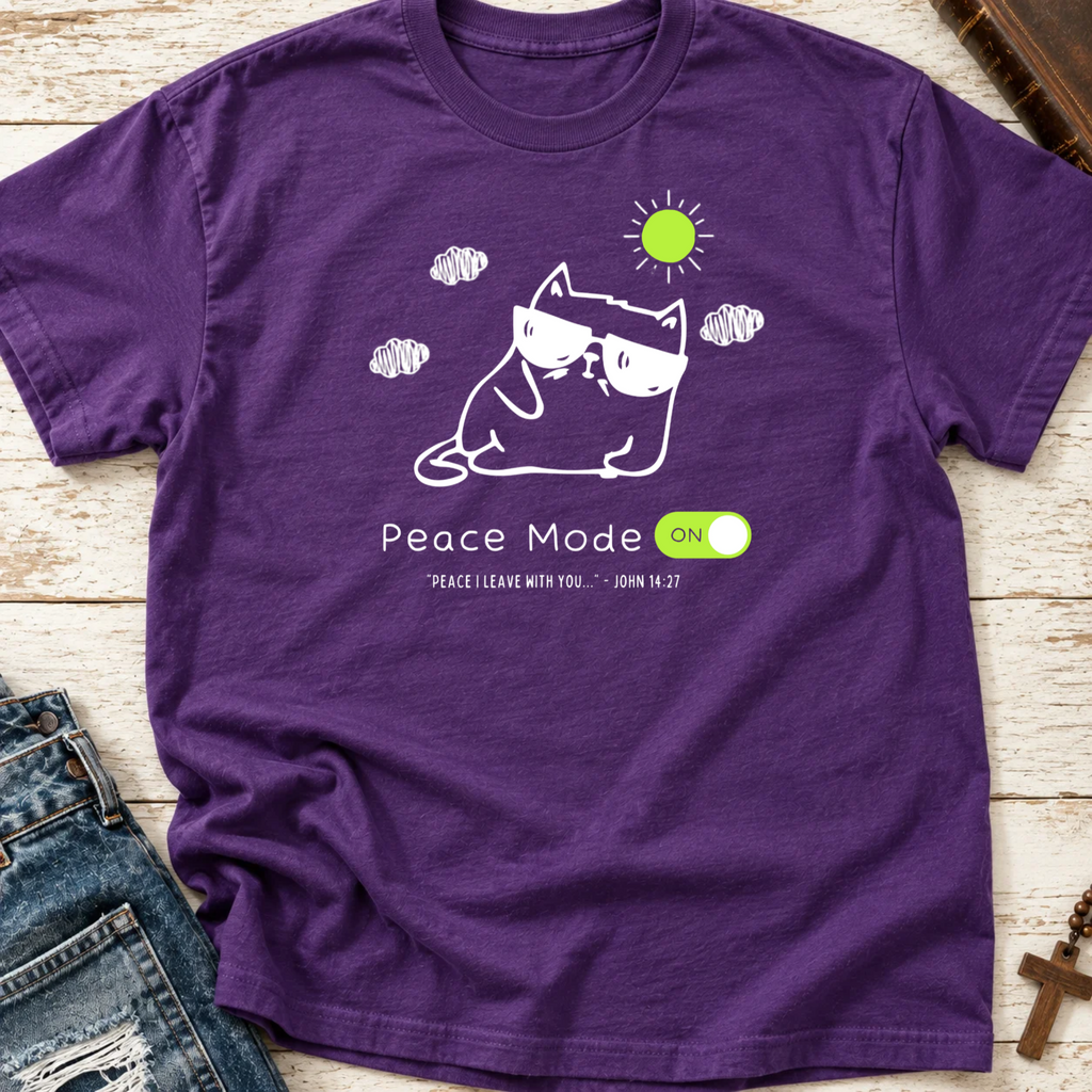 A purple graphic t-shirt laid flat on a light wood background, styled with a pair of blue jeans. The t-shirt features a white line art illustration of a cat wearing sunglasses, a green sun, and the text 'Peace Mode ON' with 'PEACE I LEAVE WITH YOU - JOHN 14:27' underneath.