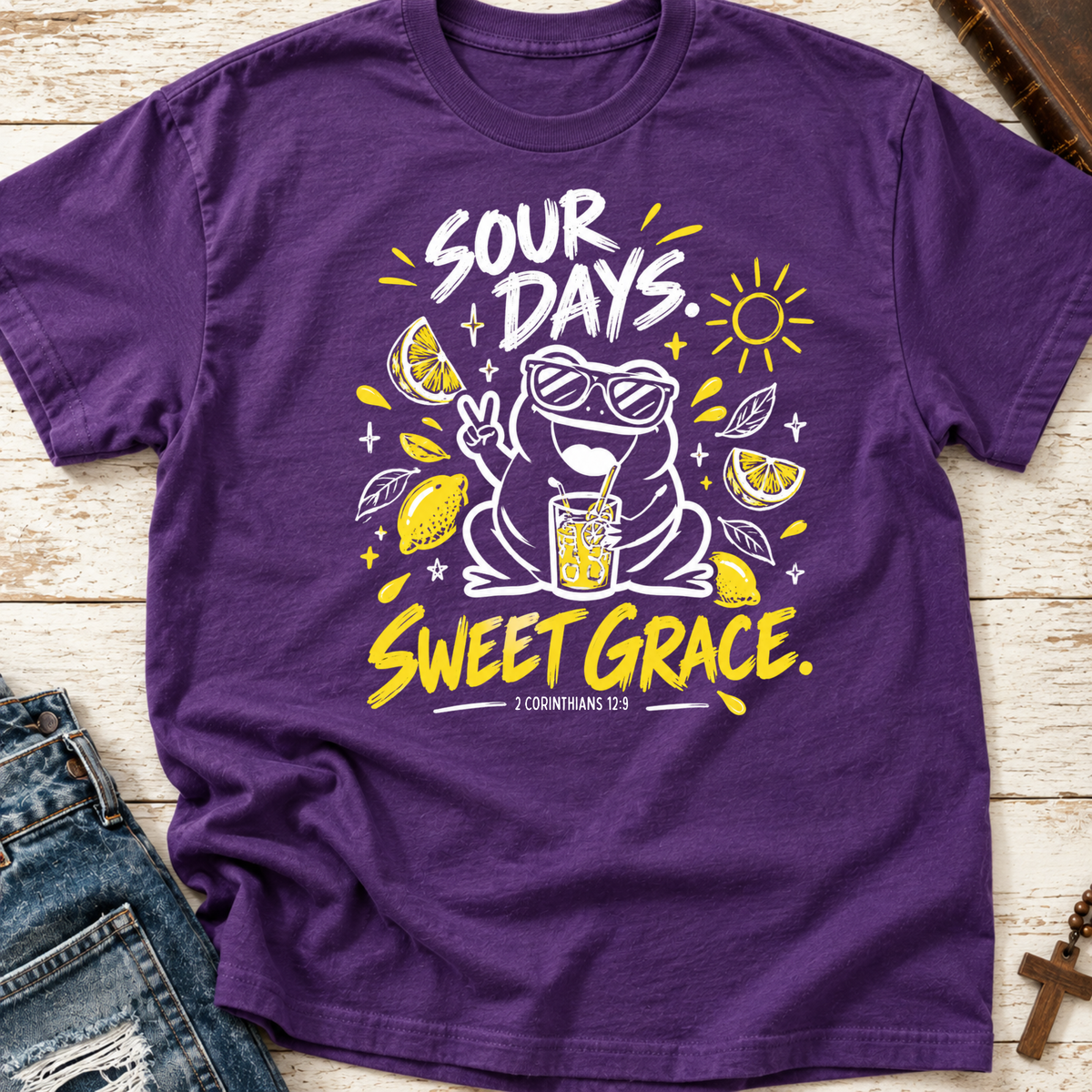 A purple short-sleeve graphic T-shirt laid flat on a light wood surface, styled with part of a blue denim jacket at the lower left. The shirt features a large front print with the words “SOUR DAYS.