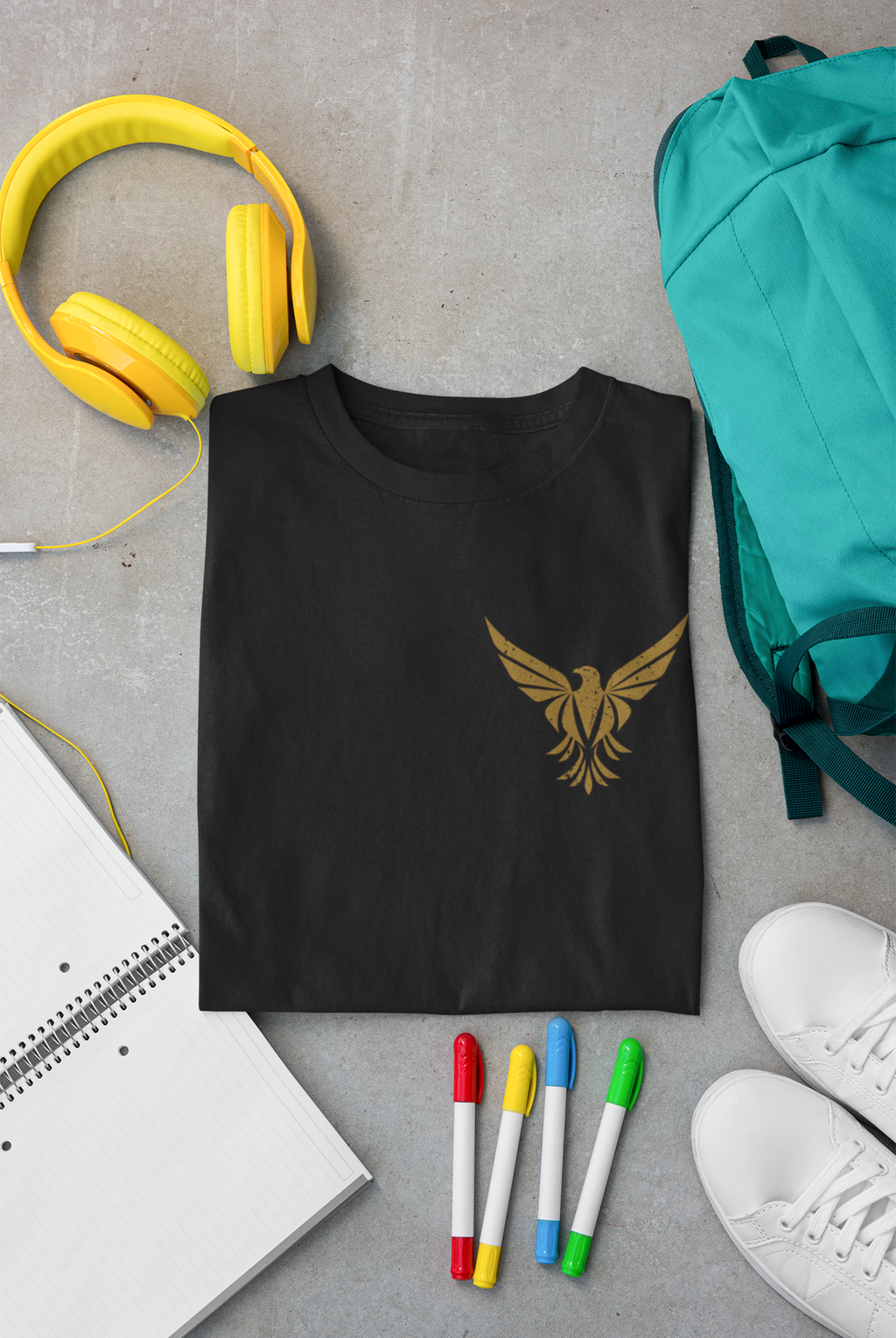 A black short-sleeve Christian graphic T-shirt is laid flat on a gray concrete surface, styled with a gold winged emblem on the left chest. Surrounding props include yellow headphones, a teal backpack, white sneakers, colored markers, and an open notebook.
