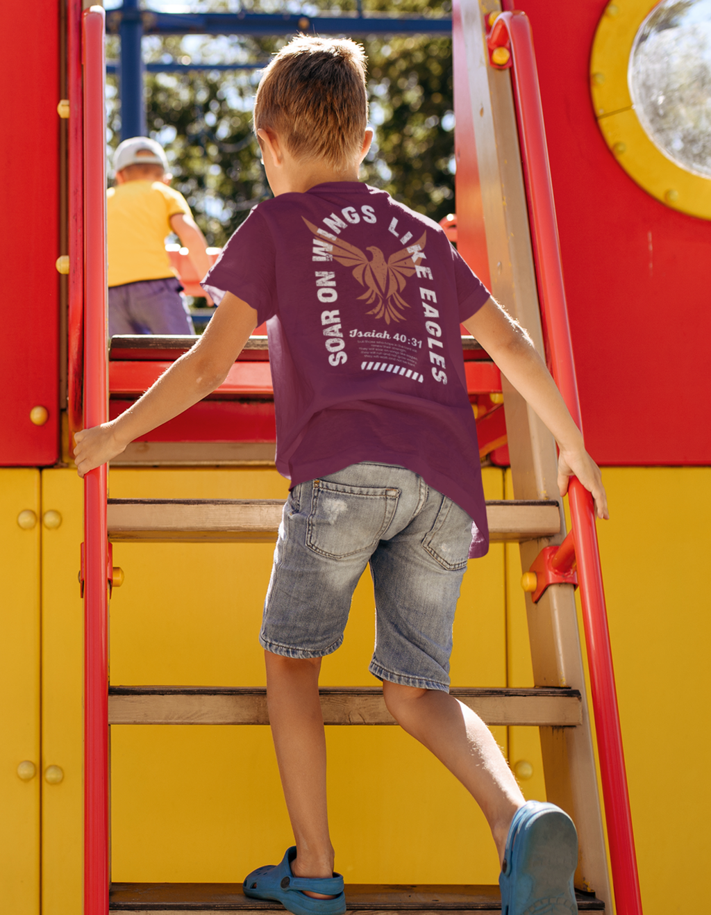 A child wearing a maroon Christian graphic t-shirt with a gold eagle graphic and the text 'SOAR ON WINGS LIKE EAGLES' climbs the red and yellow stairs of a playground structure.