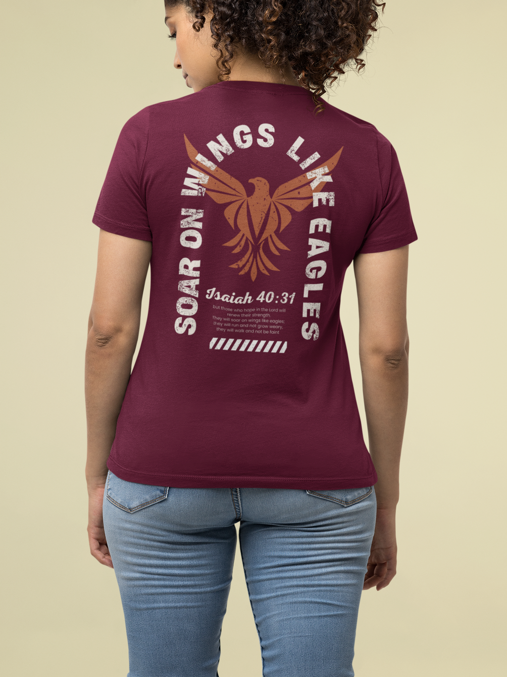 A woman viewed from the back wearing a maroon short-sleeve Christianity T-shirt with a large tan and white eagle graphic and text reading 'Soar on Wings Like Eagles' and 'Isaiah 40:31'. She is also wearing light blue jeans against a plain beige studio background.