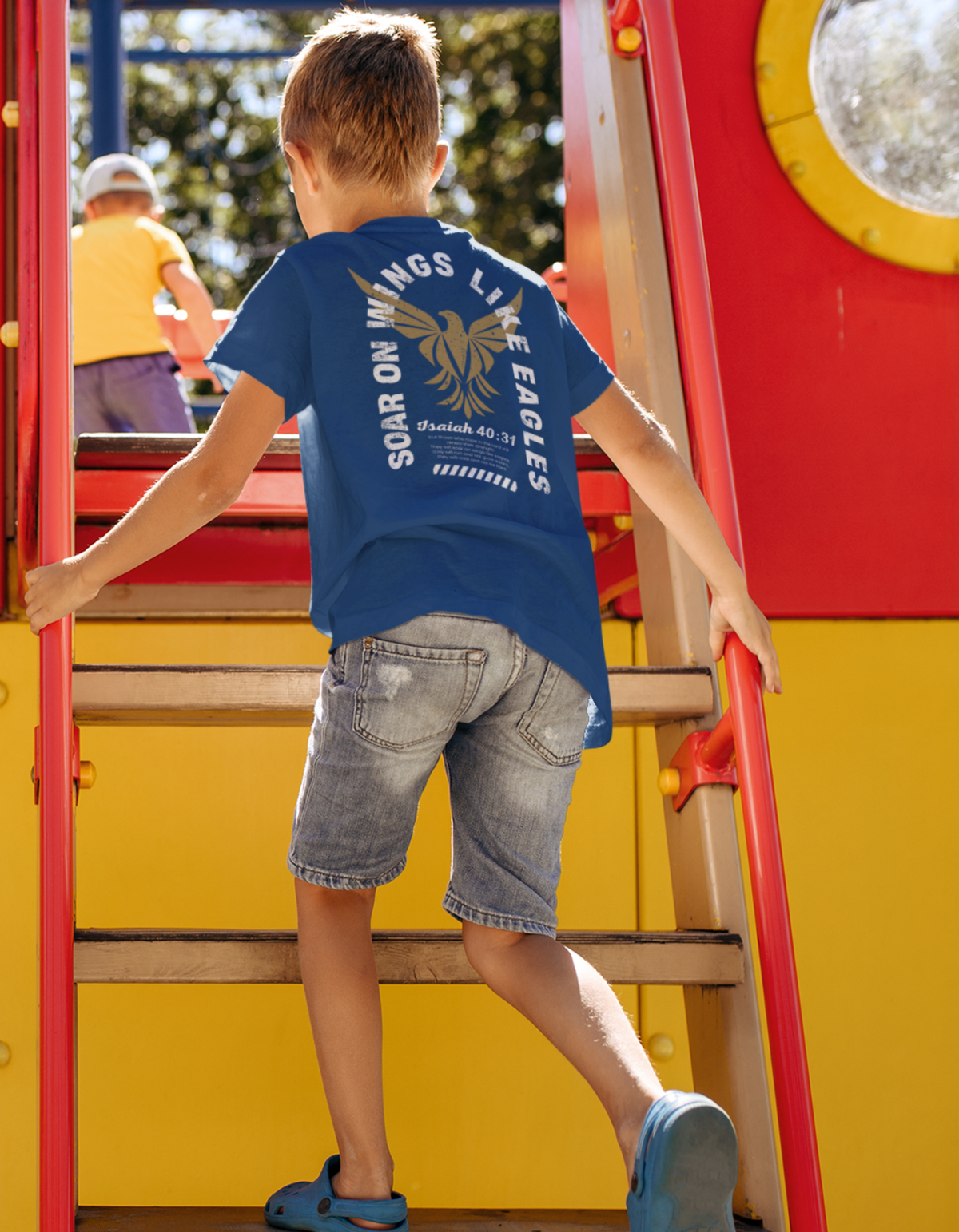 A young boy wearing a blue Christian graphic t-shirt with a gold eagle graphic and the words 'SOAR ON WINGS LIKE EAGLES' and 'ISAIAH 40:31' climbs the red and yellow playground ladder.