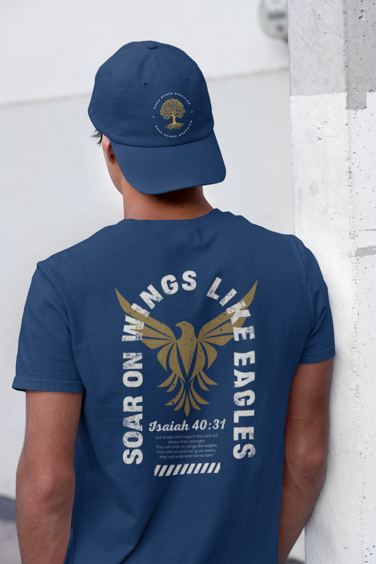 A man viewed from behind wearing a navy blue Christian graphic t-shirt and matching navy blue baseball cap. The t-shirt features a large gold eagle graphic with the words 'SOAR ON WINGS LIKE EAGLES' and 'Isaiah 40:31' printed below.