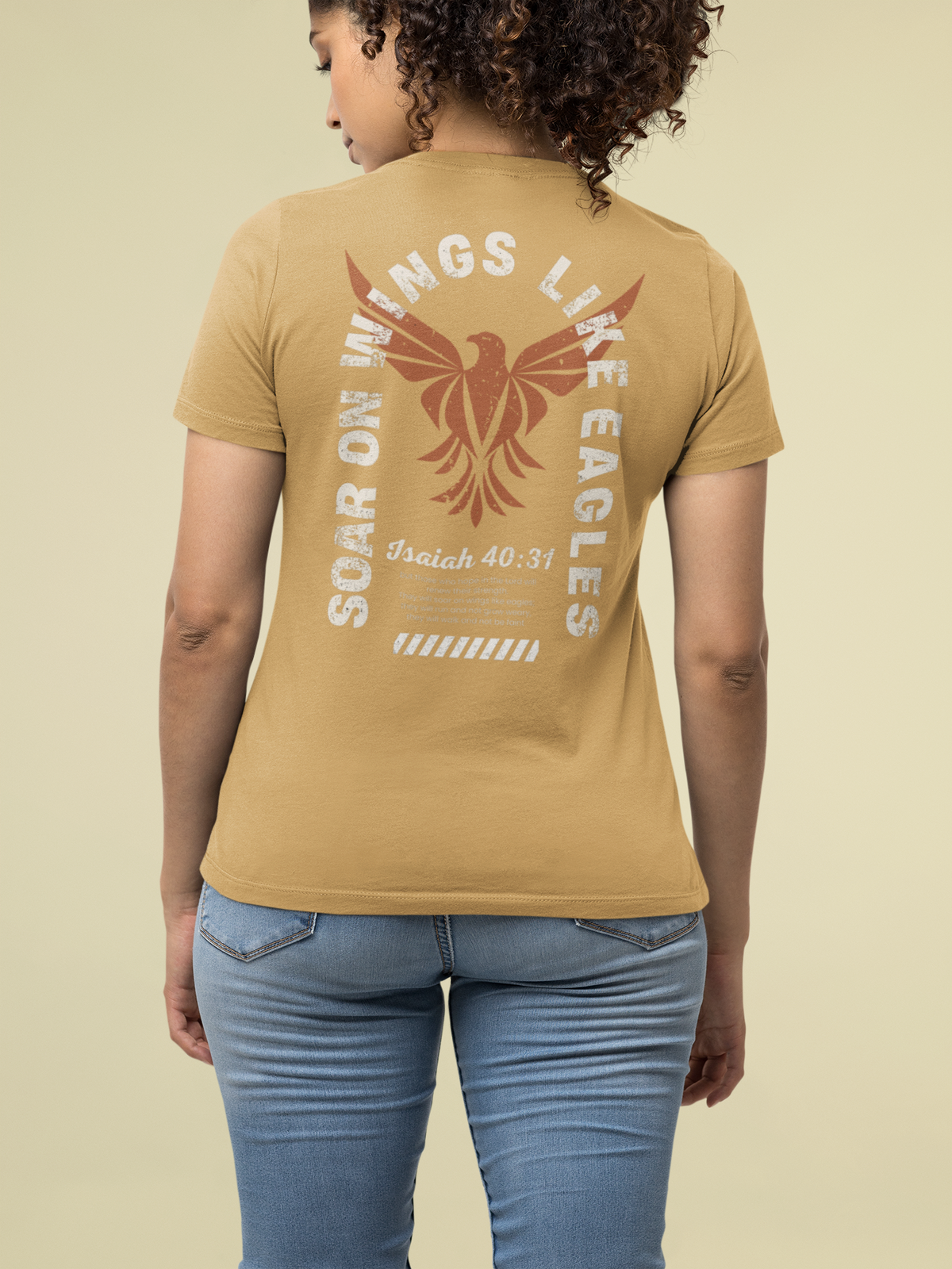 A woman shown from the back wearing a tan Christian graphic t-shirt with a large eagle design and text reading 'SOAR ON WINGS LIKE EAGLES' and 'Isaiah 40:31'. She is also wearing blue jeans against a solid light beige background.
