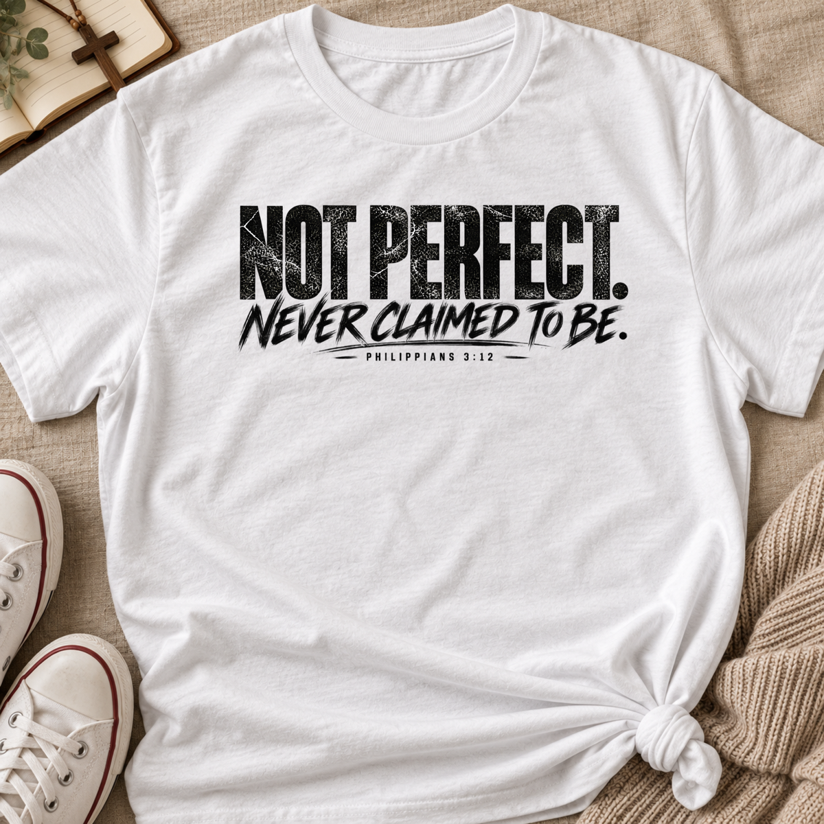 White short-sleeve T-shirt laid flat on a beige fabric surface, with distressed black text reading “NOT PERFECT. NEVER CLAIMED TO BE.